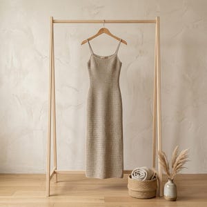 May include: A beige midi-length dress with a square neckline and thin spaghetti straps, crafted from a textured knit fabric. The garment hangs on a wooden hanger suspended from a light-colored wooden clothing rack frame.