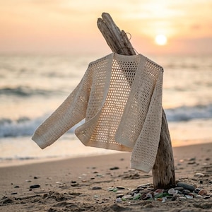 Może przedstawiać: Kremowy kardigan z otwartym splotem wisi na kawałku drewna wyrzuconego na brzeg na piaszczystej plaży. Sweter z długim rękawem wykonany jest z lekkiego, przewiewnego materiału. W tle widać ocean i zachód słońca.