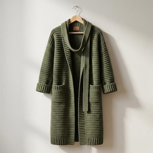 May include: An olive green, long-sleeved, open-front cardigan featuring a ribbed knit texture, a shawl collar, two front patch pockets, and a matching tie belt. The garment is displayed on a wooden hanger against a plain, light-colored wall.