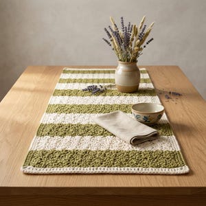 Puede incluir: Un camino de mesa rectangular de ganchillo con rayas horizontales alternas en verde oliva y crema. El camino de mesa está decorado con un jarrón de cerámica lleno de lavanda seca, un pequeño cuenco de cerámica con un patrón floral y una servilleta de lino doblada.