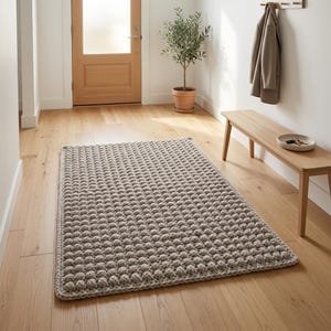 May include: A rectangular area rug featuring a thick, chunky knit texture with a repeating bobble or popcorn stitch pattern in a neutral light beige colour. The rug has a finished, braided-style border edge and lies flat on a wooden floor.