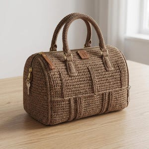 May include: A brown crocheted handbag featuring a structured rectangular shape, two top handles, and a gold-toned zipper closure. The bag includes a small leather-like patch with the letter B embossed on the front and a metal key attached to the side.