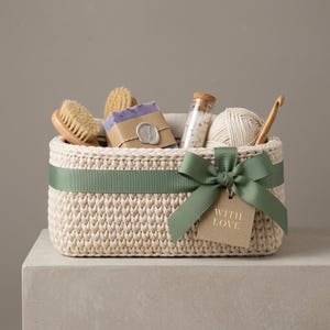 May include: A cream-colored, crocheted basket tied with a green ribbon and bow, containing a wooden brush, soap, a small glass bottle, and yarn. A tag with the words "WITH LOVE" hangs from the bow. The basket sits on a neutral surface.