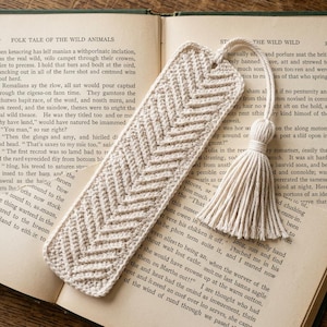 Op de afbeelding: Een crèmekleurige gehaakte boekenlegger met een gestructureerd visgraatpatroon. De boekenlegger is rechthoekig met een afgeronde bovenkant en bevat een bijpassend crèmekleurig garenkwastje dat met een lus is bevestigd.