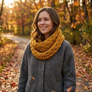May include: A woman wearing a mustard yellow crocheted cowl scarf and a gray coat. The scarf has an open, textured design. The background features a path through a forest with autumn foliage.