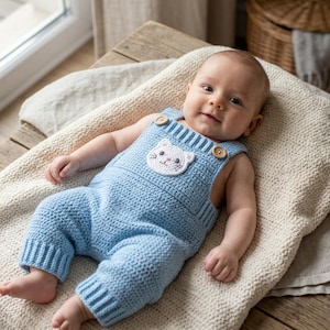 May include: A baby wearing a light blue crochet romper with a white cat applique on the chest. The romper has wooden button closures and ribbed cuffs. The baby is lying on a cream-colored blanket.
