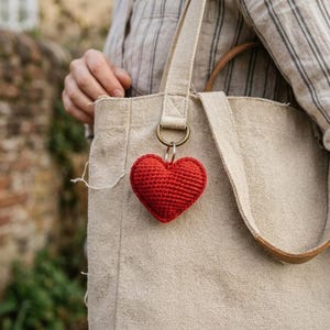 May include: A bright red, three-dimensional crocheted heart charm attached to a metal ring. The heart is suspended from the strap of a beige, textured canvas tote bag. The fabric of the bag is a neutral, light tan colour with a visible woven texture.