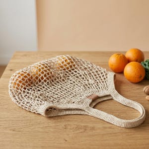 May include: A beige, crocheted reusable shopping bag filled with oranges, resting on a wooden surface. The bag has a rectangular shape with a handle. Three oranges and some greenery are next to the bag.