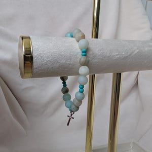 May include: A beaded bracelet with a small cross charm. The bracelet features a mix of light blue, turquoise, and neutral-toned round beads. The cross charm is silver-coloured. The bracelet is displayed on a gold jewellery stand.