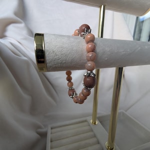 May include: A bracelet featuring peach-toned beads and dark brown feature beads. The bracelet is displayed on a white jewellery stand with gold detailing. The bracelet has silver-coloured decorative elements.