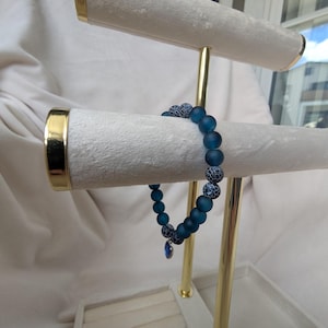 May include: A blue beaded bracelet with a small blue pendant is displayed on a white jewelry stand with gold accents. The bracelet features matte blue beads and patterned beads. The stand has a velvet-like texture.
