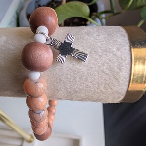 May include: A beaded bracelet featuring alternating brown wooden beads, white beads, and orange-toned stone beads. A silver-colored cross charm hangs from the bracelet. The bracelet is displayed on a light-colored, textured surface.