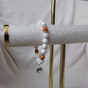 May include: A beaded bracelet with white and orange beads, a silver angel charm, and a pearl. The bracelet has cross-decorated orange beads. The bracelet is displayed on a gold jewellery stand.