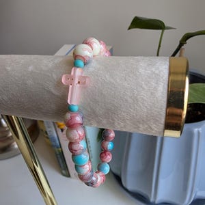 May include: A bracelet with pink, white, and turquoise beads, and a pink cross charm. The bracelet is displayed on a white surface. The background includes a gold-coloured metal stand and a potted plant.