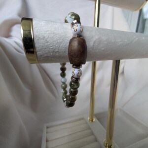 May include: A beaded bracelet with a dark brown wooden barrel bead and olive green and white beads. The bracelet is displayed on a jewellery stand with gold accents. The beads are strung on a stretchy cord.