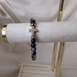 May include: A bracelet with a silver cross charm and black and clear beads. The bracelet is displayed on a white, velvet-covered jewellery stand with gold accents. The beads are approximately 0.8 cm in diameter.