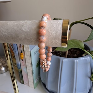 May include: Two beaded bracelets. One bracelet features peach-coloured beads and a gold cross charm. The other bracelet has peach and white beads. The bracelets are displayed on a white, cylindrical jewellery holder. A potted plant and books are in the background.