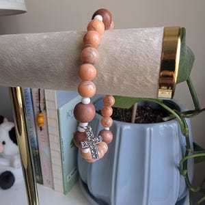 May include: A beaded bracelet with a cross charm. The bracelet features a mix of round, peach-coloured and brown beads. The cross charm is silver-coloured and encrusted with small, clear stones. The bracelet is displayed on a light-coloured, cylindrical stand.