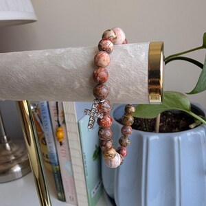 May include: A beaded bracelet with a cross charm. The bracelet has round, speckled beads in brown, red, and cream. A silver-coloured cross charm with clear stones hangs from the bracelet. The bracelet is displayed on a white surface.