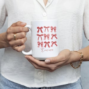 Puede incluir: Taza de cerámica blanca con un diseño de lazos rojos de varios estilos. El nombre "Emma" está escrito en cursiva debajo de los lazos. La taza es sostenida por una persona que lleva una camisa blanca y vaqueros azules.