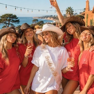 Puede incluir: Un grupo de cinco personas con camisetas rojas y sombreros de vaquero marrones, con una persona en el centro que lleva una camiseta blanca y una banda rosa con el texto Bride to Be impreso en oro, todas sosteniendo copas de vino al aire libre.