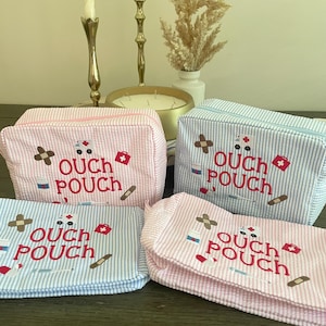 May include: Four seersucker "Ouch Pouch" cosmetic bags in pink and blue with white stripes. Each bag features the text "Ouch Pouch" and medical-themed graphics. The bags are zippered and rectangular.