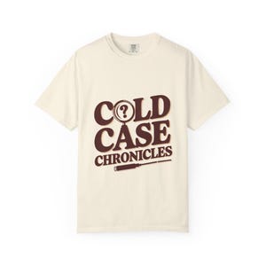 May include: A cream-colored short-sleeve t-shirt featuring the dark brown text COLD CASE CHRONICLES. The letter O in COLD is replaced by a magnifying glass with a question mark inside, and a screwdriver graphic is positioned below the text.