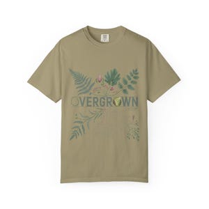 May include: A sage green short-sleeve t-shirt featuring a botanical illustration of ferns and foliage surrounding the text OVERGROWN in a dark grey font. The graphic includes various green leaves, small pink buds, and root structures.