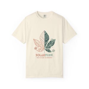 May include: Cream-colored t-shirt featuring a leaf design with circuit board patterns in red, green, and white. The text "SOLARPUNK THE FUTURE IS ORGANIC" is printed below the leaf. The shirt has a crew neck and short sleeves.