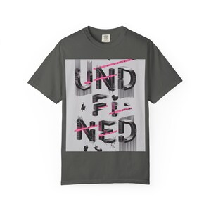 May include: Dark gray t-shirt with the word "UNDEFINED" in a 3D design on the front. The word is black with pink accents and is set against a gray background. The shirt is a casual wear item.