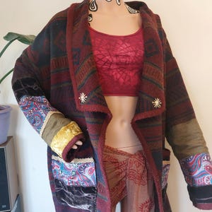 May include: A long, patterned coat in shades of burgundy, navy, and olive green. The coat features a patchwork design on the sleeves and pockets, with gold star-shaped buttons. The coat is displayed on a mannequin.