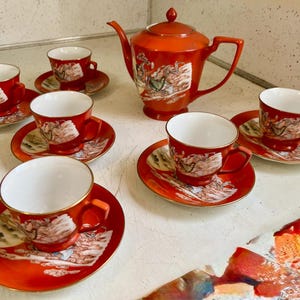 May include: A vintage tea set in a vibrant orange hue, featuring a teapot, six teacups, and saucers. Each piece is adorned with a detailed landscape design and gold trim, creating an elegant and classic aesthetic. The set is ideal for serving tea.