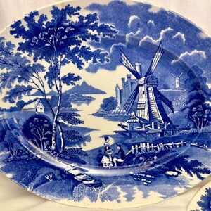 May include: A blue and white ceramic plate with a detailed landscape scene, including a windmill, trees, and figures. The plate has a vintage feel, with a classic design in a traditional style, reminiscent of antique tableware.