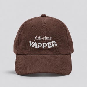 May include: A dark brown corduroy baseball cap featuring the embroidered text full-time YAPPER in white. The cap has a structured crown, a curved brim, and a button on top, showcasing a textured fabric finish with visible vertical corduroy lines.