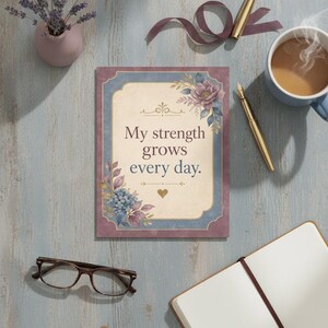 Puede incluir: Una tarjeta rectangular con un centro de color crema y el texto My strength grows every day. La tarjeta está enmarcada por un borde azul y morado decorado con ilustraciones florales en tonos azules, morados y dorados, incluyendo un pequeño corazón dorado.