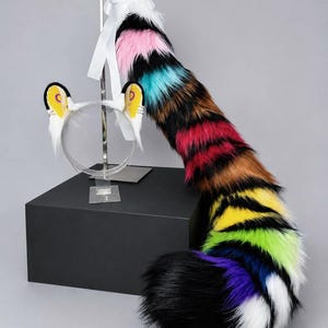May include: A set of faux fur animal ears on a headband and a long, multi-colored faux fur tail. The ears are white with black edges and yellow inner details. The tail features horizontal stripes of pink, black, teal, brown, red, yellow, lime green, blue, purple, and white.