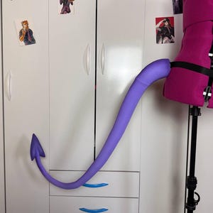 Könnte beinhalten: Ein langer, glatter, violetter Teufelsschwanz mit einer pfeilförmigen Spitze. Der Schwanz ist an einem schwarzen, verstellbaren Gurt befestigt, der um eine leuchtend pinke Torso-Schaufensterpuppe gelegt ist und sich in einer Kurve nach außen erstreckt.