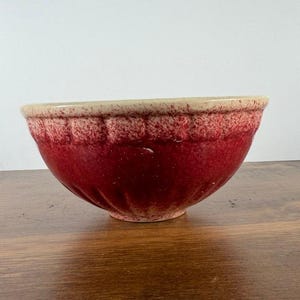 May include: A round ceramic mixing bowl featuring a deep red base color that transitions into a lighter cream-colored rim. The exterior surface displays a textured, speckled finish with vertical fluted indentations around the upper section.
