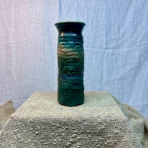 May include: A cylindrical ceramic vase featuring a textured, rugged surface with a gradient of deep teal, forest green, and earthy brown tones. The vessel has a smooth, flared rim and a solid, flat base, showcasing a handcrafted, uneven finish.
