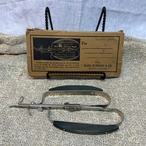May include: A vintage metal rug needle tool with black handles and a matching cardboard box labeled Sears, Roebuck & Co. The box features printed text including 'Best Rug Needle Made' and 'Make A Rug in a Day' with instructions for threading.