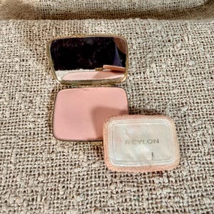 May include: A rectangular gold-tone metal compact case with an open lid revealing a mirror and a light pink powder cake. A separate rectangular powder puff with a white surface and the text REVLON printed in black is positioned next to the compact.