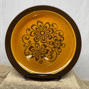 Puede incluir: Un plato redondo de cerámica con un centro de color amarillo mostaza moteado, un patrón floral marrón oscuro y un borde marrón oscuro sólido. El diseño incluye tres flores estilizadas rodeadas de acentos en forma de enredadera.