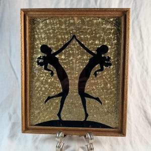 May include: A framed wall art piece featuring two black silhouettes of dancing figures against a textured gold foil background. The figures are mirrored, touching hands at the top, set within a rectangular wooden frame with ornate detailing.
