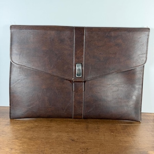 May include: A dark brown document portfolio with a textured finish, featuring a horizontal flap closure secured by a vertical strap and a rectangular silver-toned metal buckle centered on the front of the rectangular case.