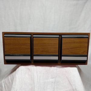 May include: A rectangular wall-mounted storage unit featuring three individual compartments. Each compartment has a wood-grain finish front panel, a black frame, and a horizontal silver-toned accent strip along the bottom edge of the drawer.