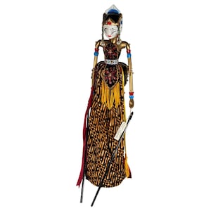 May include: A Wayang Golek rod puppet featuring a white face with painted features, a gold-toned bodice with red floral accents, and a long skirt with a brown and black batik pattern. The puppet has articulated arms with black control rods.
