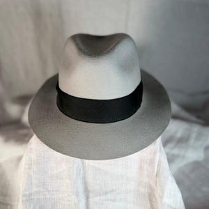 Puede incluir: Un sombrero fedora de color gris claro con una copa estructurada con pliegue central y un ala ancha y plana. El sombrero está acentuado por una cinta de grosgrain negra sólida que rodea la base de la copa, creando un contraste marcado.