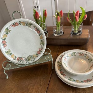 May include: A set of ceramic plates and bowls with a fruit design border. A metal serving stand holds a large plate. Red tulips in glass vases sit on a wooden tray. The table is made of wood.