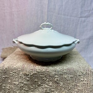 May include: A white ceramic tureen with a matching lid, featuring a scalloped edge and a decorative handle on top. The bowl has two side handles and a pedestal base, showcasing a smooth, solid white finish with subtle embossed detailing.