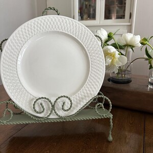 Puede incluir: Un plato de cerámica blanca con un patrón de tejido de cesta texturizado en el borde, exhibido verticalmente sobre un soporte de metal verde salvia con detalles de alambre trenzado. El plato tiene un centro liso y un borde decorativo.